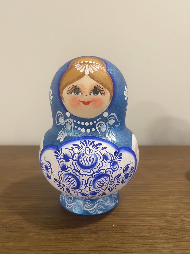 Pu&ograve; includere: Una matrioska in legno blu con un volto dipinto a mano, capelli castani, un copricapo floreale bianco e labbra rosse. La sezione inferiore presenta intricati motivi floreali blu e bianchi con delicati ghirigori e punti bianchi.