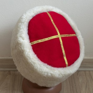 Könnte beinhalten: Ein Winterhut mit roter Oberseite und weißem Fellbesatz. Die rote Oberseite ist mit goldenem Band in einem Kreuzmuster verziert. Der Hut wird auf einem braunen Schaufensterkopf präsentiert.