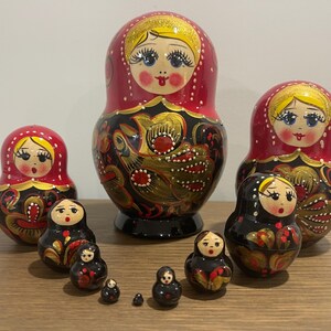 K&ouml;nnte beinhalten: Eine Sammlung russischer Matrjoschka-Puppen in verschiedenen Gr&ouml;&szlig;en. Die Puppen haben ein rot-schwarzes Farbschema mit goldenen Akzenten und bemalten Gesichtern. Die gr&ouml;&szlig;te Puppe ist etwa 15 cm hoch.