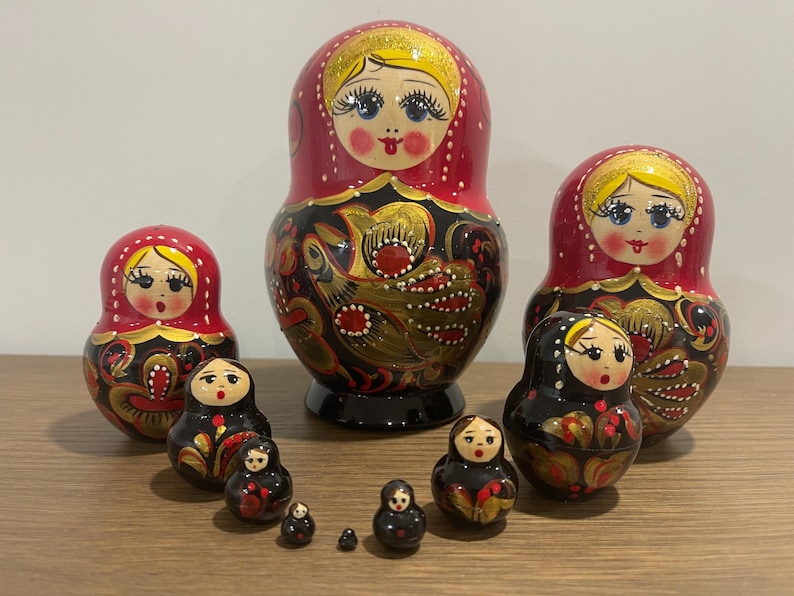 K&ouml;nnte beinhalten: Eine Sammlung russischer Matrjoschka-Puppen in verschiedenen Gr&ouml;&szlig;en. Die Puppen haben ein rot-schwarzes Farbschema mit goldenen Akzenten und bemalten Gesichtern. Die gr&ouml;&szlig;te Puppe ist etwa 15 cm hoch.