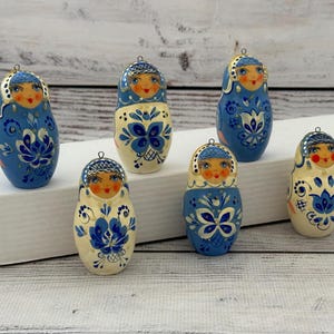 May include: Six hand-painted Matryoshka dolls with a blue and white floral design and painted faces. Each doll has a small metal loop at the top. The dolls are displayed on a white cardboard box. The dolls are a traditional Russian craft.