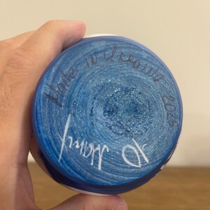 Pu&ograve; includere: La base circolare di un oggetto in ceramica presenta una superficie dipinta di blu vibrante con una consistenza glitterata. Un testo scritto a mano in inchiostro scuro recita Made in Ukraine 2025, con una firma bianca nella parte inferiore.