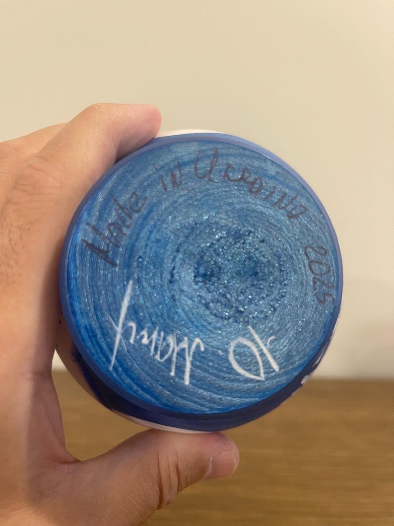 Pu&ograve; includere: La base circolare di un oggetto in ceramica presenta una superficie dipinta di blu vibrante con una consistenza glitterata. Un testo scritto a mano in inchiostro scuro recita Made in Ukraine 2025, con una firma bianca nella parte inferiore.