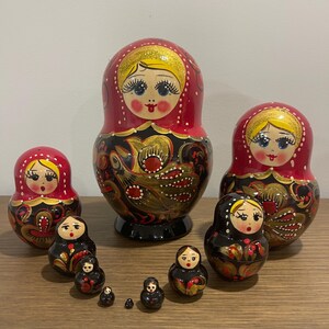 K&ouml;nnte beinhalten: Eine Sammlung russischer Matrjoschka-Puppen in verschiedenen Gr&ouml;&szlig;en. Die Puppen haben ein rot-schwarzes Farbschema mit goldenen Akzenten und bemalten Gesichtern. Die gr&ouml;&szlig;te Puppe ist etwa 15 cm hoch.