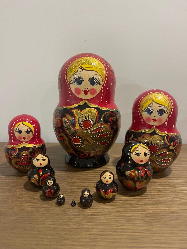 K&ouml;nnte beinhalten: Eine Sammlung russischer Matrjoschka-Puppen in verschiedenen Gr&ouml;&szlig;en. Die Puppen haben ein rot-schwarzes Farbschema mit goldenen Akzenten und bemalten Gesichtern. Die gr&ouml;&szlig;te Puppe ist etwa 15 cm hoch.