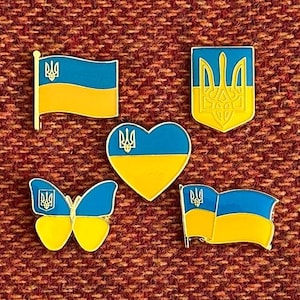 Può includere: Cinque spille con il design della bandiera ucraina in diverse forme. Le spille sono gialle e blu con un simbolo di tridente. Le spille sono su uno sfondo di tessuto rosso.