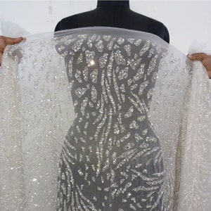 May include: White mesh fabric with a silver glitter pattern. The fabric has a geometric design with a repeating pattern of small, silver sequins. The fabric is sheer and has a soft, flowing texture. The fabric is labeled "TUCF The Urban Couture Factory".
