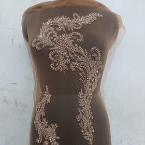 May include: A gold beaded applique with a floral design on a brown mesh fabric. The applique is perfect for adding a touch of elegance to any garment.