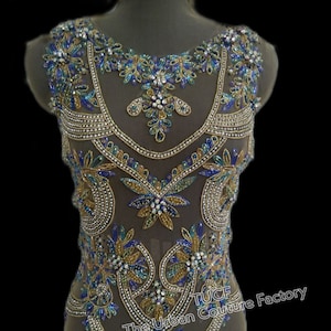 Op de afbeelding: Een doorschijnend, mouwloos topje versierd met gouden en blauwe kralen en strass. Het topje heeft een bloemenpatroon en de tekst "The Urban Couture Factory" is zichtbaar aan de onderkant.