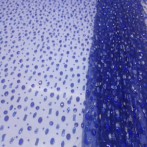 May include: Close-up of a sheer fabric embellished with blue and silver-toned rhinestones. The rhinestones are in various shapes and sizes, creating a sparkling effect. The fabric is a deep blue color.