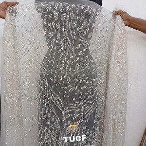 May include: White sheer fabric with a silver glitter pattern. The pattern is a series of abstract shapes and lines. The fabric is draped over a dress form. The text "TUCF The Urban Couture Factory" is printed on the fabric.