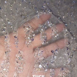 May include: Close-up of a sheer, white fabric with silver sequins and beads. The fabric has a delicate, sparkly texture. The text "TUCF The Urban Couture Factory" is visible in the bottom right corner.