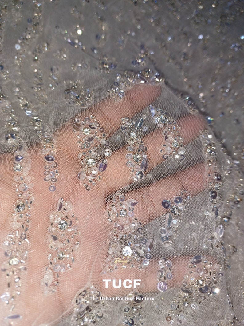 May include: Close-up of a sheer, white fabric with silver sequins and beads. The fabric has a delicate, sparkly texture. The text "TUCF The Urban Couture Factory" is visible in the bottom right corner.