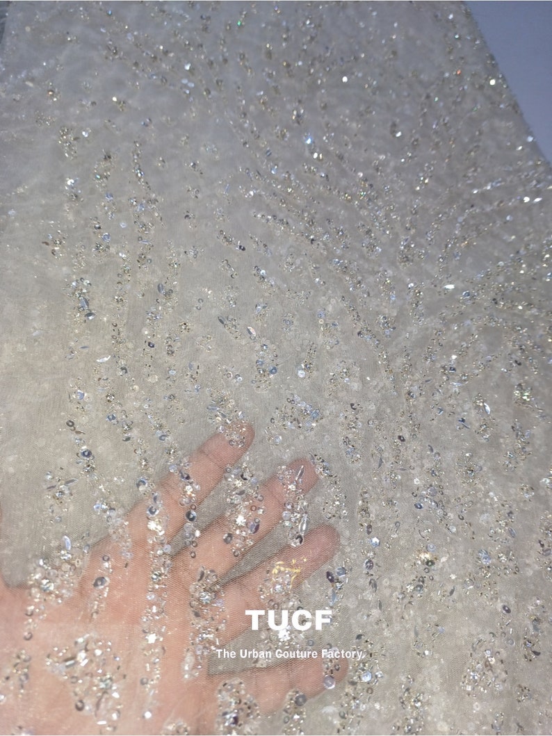 May include: White sheer fabric with a scattered pattern of small, silver sequins. The fabric has a delicate, sparkly appearance. The text "TUCF The Urban Couture Factory" is printed in the bottom right corner.