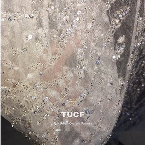 May include: Close-up of a sheer white fabric with silver sequins and beads. The fabric has a delicate, intricate pattern. The text "TUCF The Urban Couture Factory" is visible in the lower right corner.