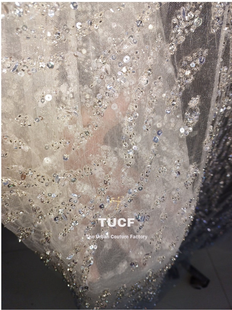 May include: Close-up of a sheer white fabric with silver sequins and beads. The fabric has a delicate, intricate pattern. The text "TUCF The Urban Couture Factory" is visible in the lower right corner.