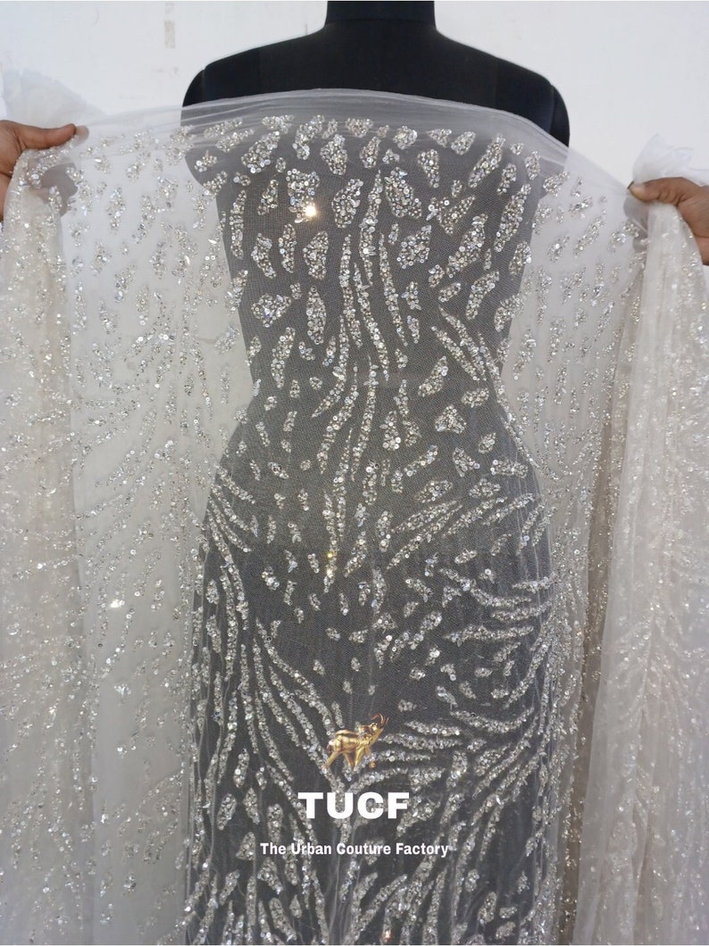 May include: White mesh fabric with a silver glitter pattern. The fabric has a geometric design with a repeating pattern of small, silver sequins. The fabric is sheer and has a soft, flowing texture. The fabric is labeled "TUCF The Urban Couture Factory".