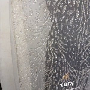 May include: Close-up of a white sheer fabric with a silver glitter pattern. The fabric has a delicate, flowing texture and is adorned with a repeating design of small, shimmering sequins. The fabric is perfect for creating elegant and glamorous garments.