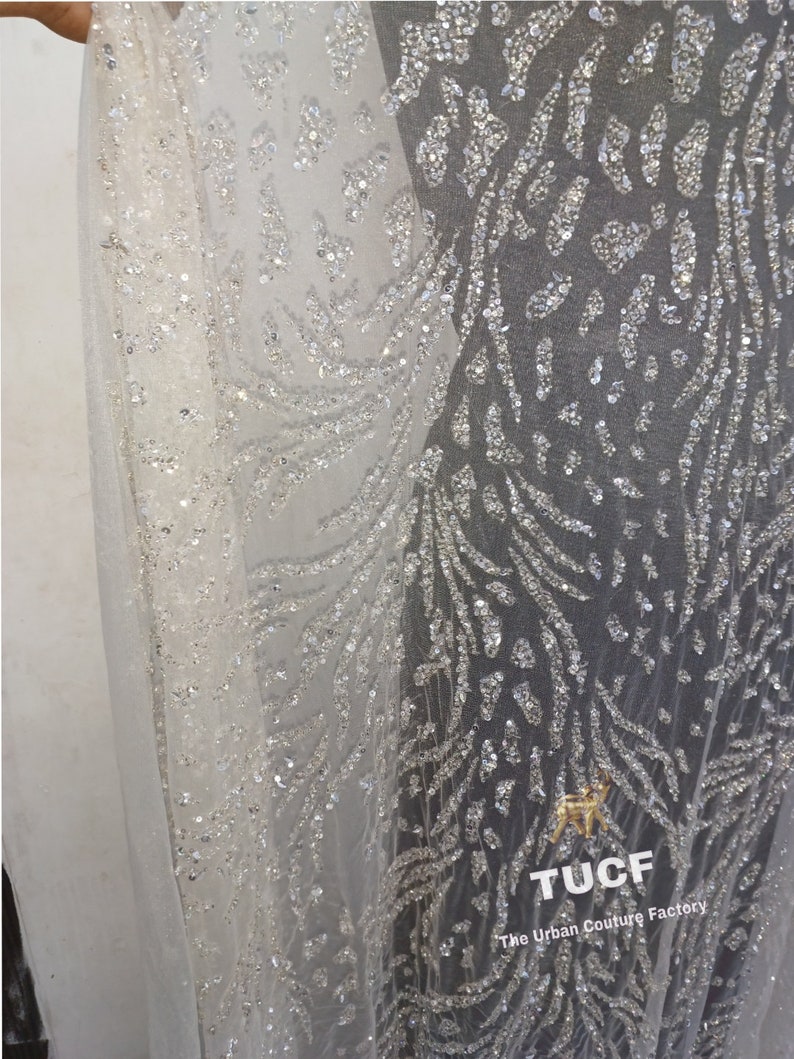 May include: Close-up of a white sheer fabric with a silver glitter pattern. The fabric has a delicate, flowing texture and is adorned with a repeating design of small, shimmering sequins. The fabric is perfect for creating elegant and glamorous garments.