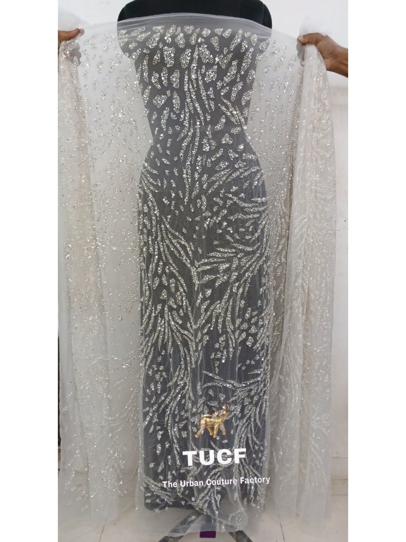 May include: White sheer fabric with silver glitter and a silver leaf pattern. The fabric is draped over a mannequin. The text "TUCF The Urban Couture Factory" is printed at the bottom of the image.