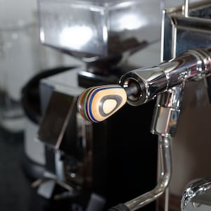 Puede incluir: Primer plano de una máquina de espresso cromada. La máquina tiene un mango de madera multicapa y colorido. Un manómetro es visible a la derecha. El fondo está borroso, lo que sugiere un entorno de cocina.