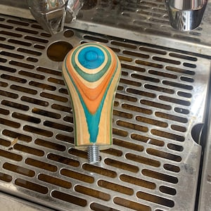May include: A colorful wooden espresso tamper with a blue, green, orange, and brown swirl pattern. The tamper is sitting on a stainless steel drip tray.
