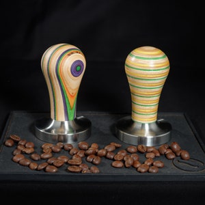 Könnte beinhalten: Zwei Kaffeettamper aus Holz mit verschiedenen bunten Mustern auf einer schwarzen Oberfläche mit Kaffeebohnen, die um sie herum verstreut sind.