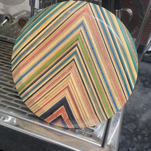 May include: A round, colorful coaster made from layers of wood. The wood is arranged in a chevron pattern with shades of brown, black, red, blue, green, and yellow.