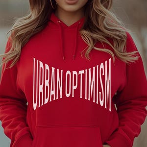 May include: A red hooded sweatshirt with the text "URBAN OPTIMISM" in white. The sweatshirt has a front pocket and is being worn with light blue jeans. The person is standing outside in front of a blurred background.