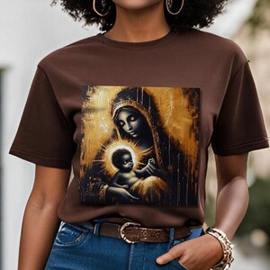 May include: A dark brown short-sleeve t-shirt featuring a square graphic print of a mother and child in a gold and black artistic style. The mother wears a head covering and the child is centered in a glowing halo, set against a dark background.