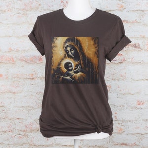 May include: A dark brown short-sleeved t-shirt featuring a central graphic print of a mother and child in gold and black tones. The artwork depicts a figure with a halo holding a baby, rendered in a painterly style with vertical drip effects.