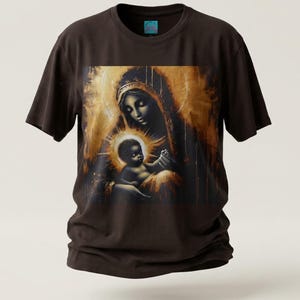 May include: A dark brown short-sleeve t-shirt featuring a central graphic print of a figure holding a child, rendered in shades of gold, bronze, and black. The artwork displays a glowing halo effect around the child and a textured, painterly style.