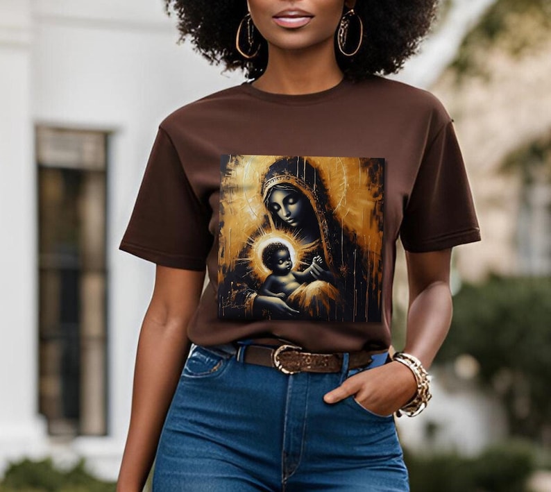 May include: A dark brown short-sleeve t-shirt featuring a square graphic print of a mother and child in a gold and black artistic style. The mother wears a head covering and the child is centered in a glowing halo, set against a dark background.