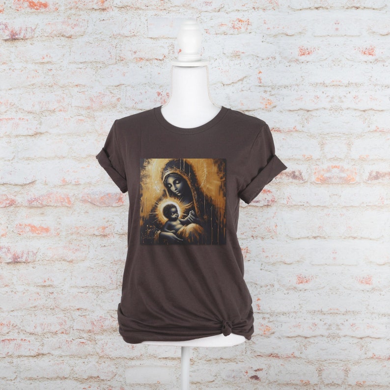 May include: A dark brown short-sleeved t-shirt featuring a central graphic print of a mother and child in gold and black tones. The artwork depicts a figure with a halo holding a baby, rendered in a painterly style with vertical drip effects.