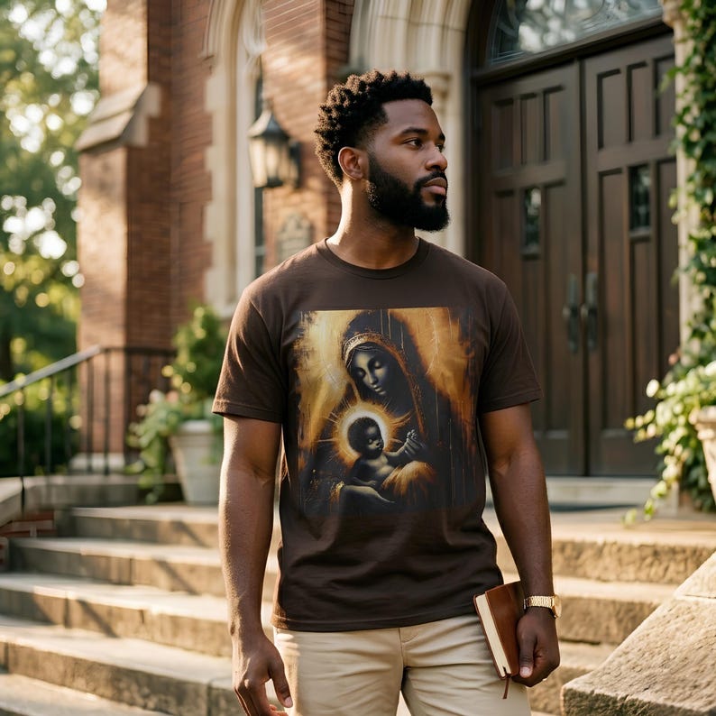 May include: A dark brown short-sleeved t-shirt featuring a central graphic print of a mother and child in a golden, painterly style. The shirt is paired with light beige trousers and a small brown book held in one hand.
