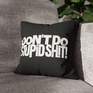 May include: A square black throw pillow featuring the white text DON'T DO STUPID SHIT! printed in a bold, sans-serif font across the center. The pillow has a smooth fabric texture and is positioned against a gray upholstered furniture surface.