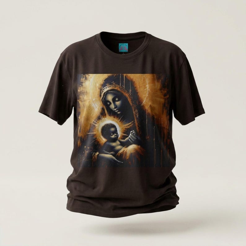 May include: A dark brown short-sleeve t-shirt featuring a central graphic print of a figure holding a child, rendered in shades of gold, bronze, and black. The artwork displays a glowing halo effect around the child and a textured, painterly style.