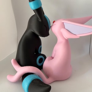 May include: Two stylised animal figurines sitting face to face. The left figure is black with bright blue circular accents and rings on its ears and tail. The right figure is light pink with large lavender-toned ears and a small red dot on its forehead.