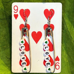 Puede incluir: Pendientes hechos a mano con imperdibles plateados y cuentas blancas que deletrean "LOVE". Cada cuenta tiene un corazón verde. Los pendientes cuelgan de ganchos plateados. Los pendientes se exhiben en una carta de juego con corazones rojos.