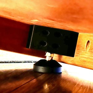 May include: Close-up of a black furniture leg with a round base and a square metal plate with holes. The leg is attached to a wooden surface.