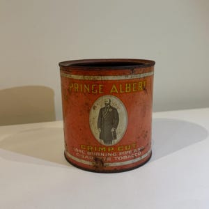 May include: A vintage Prince Albert Crimp Cut tobacco tin with a red and orange label. The label features a man in a suit and a hat, and the text "Prince Albert Crimp Cut Long Burning Pipe and Cigarette Tobacco".