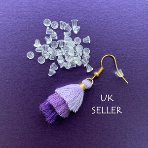 May include: A purple tassel earring with a gold hook and a clear silicone earring back. A pile of clear silicone earring backs are also shown in the image. The text "UK SELLER" is written in white on a purple background.