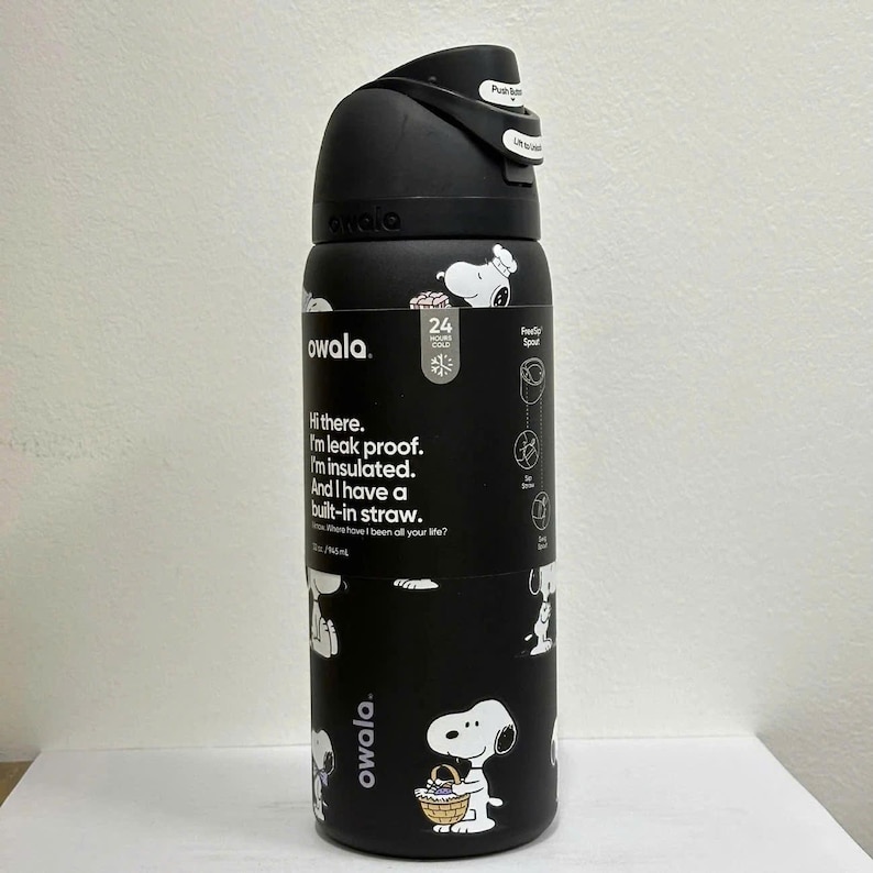 Op de afbeelding: Een zwarte Owala ge&iuml;soleerde waterfles met een Snoopy-patroon en een zwarte dop met drukknopmechanisme. De fles bevat een label met de tekst: Hi there. I'm leak proof. I'm insulated. And I have a built-in straw. 945 ml / 32 oz.