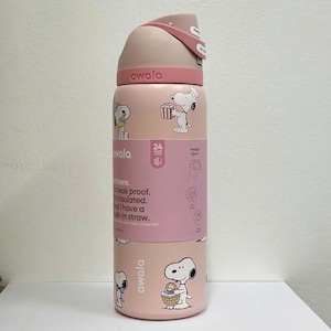 Op de afbeelding: Een lichtroze ge&iuml;soleerde Owala-waterfles met een Snoopy-print. De fles heeft een deksel met drukknop en draaglus, een ingebouwd rietje en een label met de tekst: Hi there. I'm leak proof. I'm insulated. And I have a built-in straw.