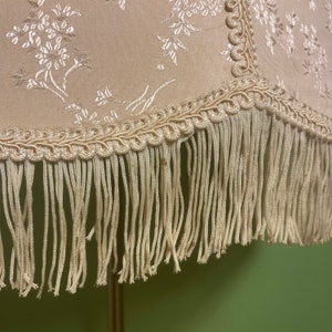 May include: A cream-colored lampshade with a floral pattern and a fringe trim. The fringe is made of a cream-colored fabric.