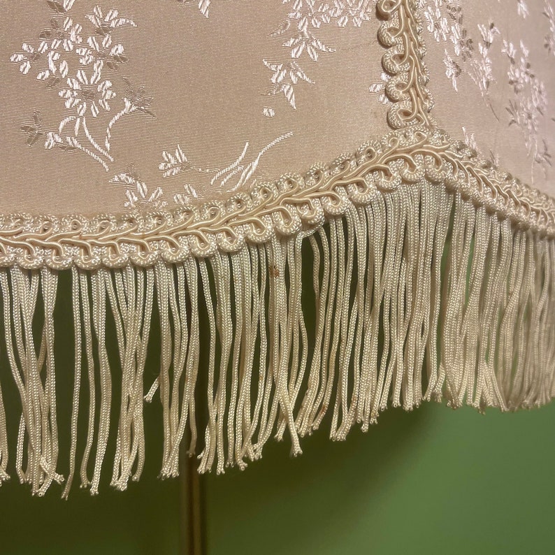 May include: A cream-colored lampshade with a floral pattern and a fringe trim. The fringe is made of a cream-colored fabric.