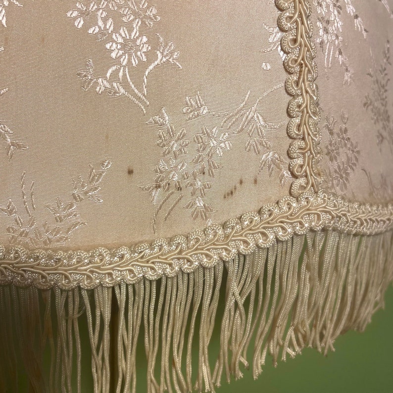 May include: A cream-colored lampshade with a floral pattern and a fringe trim. The fringe is made of a cream-colored fabric.