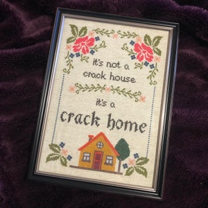 May include: Framed cross-stitch art with the text "It's not a crock house, it's a crack home." The artwork features a house, tree, and floral border in shades of red, green, and yellow. The frame is black.