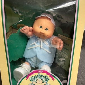 May include: A Cabbage Patch Kids Preemie doll in a blue and white outfit with a white hat. The doll is sitting in a box with a green background and the text "1985 Cabbage Patch Kids Preemie!" and "I'm unique. I'm adoptable!" The box also has a March of Dimes logo.