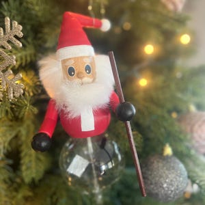 May include: A wooden Santa Claus ornament with a red hat, white beard, and a brown staff. The ornament is hanging on a Christmas tree.